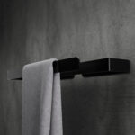 Wieszak łazienkowy w minimalistycznym czarnym matowym wykończeniu zamontowany na ciemnoszarej betonowej ścianie. Na wieszaku znajduje się elegancko przewieszony jasnoszary ręcznik. Całość tworzy nowoczesną i stylową aranżację łazienki.