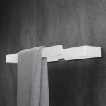 Biały, minimalistyczny wieszak na ręczniki wykonany z aluminium, zamocowany na ciemnoszarej, betonowej ścianie. Prosta, geometryczna forma wieszaka podkreśla nowoczesny design, a matowe wykończenie dodaje elegancji. Na wieszaku zawieszony jest szary ręcznik, który kontrastuje z białą powierzchnią wieszaka, tworząc harmonijną, estetyczną kompozycję.