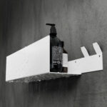 Nowoczesna, biała półka łazienkowa z haczykami, wykonana z trwałego aluminium. Konstrukcja odporna na wodę, z widocznymi kroplami na powierzchni. Na półce umieszczone są eleganckie butelki z kosmetykami. Styl loftowy, minimalistyczny design pasujący do industrialnych wnętrz.