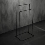 Czarny, minimalistyczny stojak łazienkowy na ręczniki AVRO ustawiony na ciemnej, betonowej podłodze przy ścianie z surowego betonu.