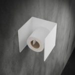 biały, metalowy uchwyt na papier toaletowy Nira, zamontowany na betonowej ścianie w nowoczesnej łazience – minimalistyczny design w stylu industrialnym