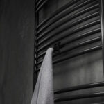 Podwójny haczyk na kaloryfer, aluminiowy, na tle eleganckiej łazienki oraz kaloryfera drabinkowego. W kolorze czarny mat