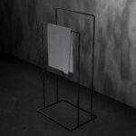 Czarny, minimalistyczny stojak łazienkowy na ręczniki AVRO ustawiony na ciemnej, betonowej podłodze przy ścianie z surowego betonu. Na stojaku zawieszony jest szary ręcznik.