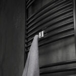 Podwójny haczyk na kaloryfer, aluminiowy, na tle eleganckiej łazienki oraz kaloryfera drabinkowego. W kolorze biały mat