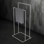 biały, minimalistyczny stojak łazienkowy na ręczniki AVRO ustawiony na ciemnej, betonowej podłodze przy ścianie z surowego betonu.