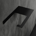 Minimalistyczny uchwyt na papier toaletowy Sora w kolorze matowej czerni, wykonany z jednego giętego elementu metalu, o nowoczesnej i geometrycznej formie – widok z ukosa na tle łazienki