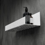 Czarna półka łazienkowa samoprzylepna z matowego metalu, pokryta kroplami wody. Na półce ustawione są dwie butelki kosmetyków – większa z pompką i mniejsza z zakrętką. Półka ma prostokątny kształt z pełnymi bokami i perforowanym spodem umożliwiającym odpływ wody.