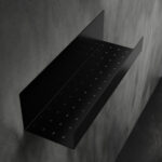 Czarna półka łazienkowa samoprzylepna z matowego metalu, pokryta kroplami wody. Na półce ustawione są dwie butelki kosmetyków – większa z pompką i mniejsza z zakrętką. Półka ma prostokątny kształt z pełnymi bokami i perforowanym spodem umożliwiającym odpływ wody.