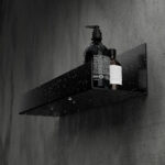 Czarna półka łazienkowa samoprzylepna z matowego metalu, pokryta kroplami wody. Na półce ustawione są dwie butelki kosmetyków – większa z pompką i mniejsza z zakrętką. Półka ma prostokątny kształt z pełnymi bokami i perforowanym spodem umożliwiającym odpływ wody.