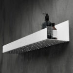 Czarna półka łazienkowa samoprzylepna z matowego metalu, pokryta kroplami wody. Na półce ustawione są dwie butelki kosmetyków – większa z pompką i mniejsza z zakrętką. Półka ma prostokątny kształt z pełnymi bokami i perforowanym spodem umożliwiającym odpływ wody.