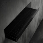 Czarna półka łazienkowa samoprzylepna z matowego metalu, pokryta kroplami wody. Na półce ustawione są dwie butelki kosmetyków – większa z pompką i mniejsza z zakrętką. Półka ma prostokątny kształt z pełnymi bokami i perforowanym spodem umożliwiającym odpływ wody.