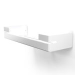 Biała, metalowa półka łazienkowa VEGA z matowym wykończeniem, zamontowana na ścianie, z widocznymi kroplami wody. Na półce ustawione są eleganckie kosmetyki w ciemnych butelkach. Konstrukcja posiada perforowane dno dla odpływu wody oraz wysokie boki zabezpieczające przed zsuwaniem się przedmiotów.