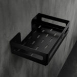 Czarna metalowa półka łazienkowa NAOS z matowym wykończeniem, zamontowana na ścianie, z perforowanym dnem i wysokimi bokami; na półce ustawione butelki kosmetyków.