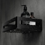 Czarna metalowa półka łazienkowa NAOS z matowym wykończeniem, zamontowana na ścianie, z perforowanym dnem i wysokimi bokami; na półce ustawione butelki kosmetyków.