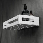 Biała metalowa półka łazienkowa NAOS z matowym wykończeniem, zamontowana na ścianie, z perforowanym dnem i wysokimi bokami; na półce ustawione butelki kosmetyków.