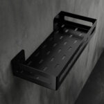 Czarna metalowa półka łazienkowa NAOS z matowym wykończeniem, zamontowana na ścianie, z perforowanym dnem i wysokimi bokami; na półce ustawione butelki kosmetyków.