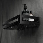 Czarna metalowa półka łazienkowa NAOS z matowym wykończeniem, zamontowana na ścianie, z perforowanym dnem i wysokimi bokami; na półce ustawione butelki kosmetyków.