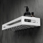 Biała metalowa półka łazienkowa NAOS z matowym wykończeniem, zamontowana na ścianie, z perforowanym dnem i wysokimi bokami; na półce ustawione butelki kosmetyków.