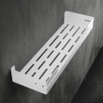 Biała, metalowa półka łazienkowa VEGA z matowym wykończeniem, zamontowana na ścianie, z widocznymi kroplami wody. Na półce ustawione są eleganckie kosmetyki w ciemnych butelkach. Konstrukcja posiada perforowane dno dla odpływu wody oraz wysokie boki zabezpieczające przed zsuwaniem się przedmiotów.