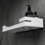 Biała, metalowa półka łazienkowa VEGA z matowym wykończeniem, zamontowana na ścianie, z widocznymi kroplami wody. Na półce ustawione są eleganckie kosmetyki w ciemnych butelkach. Konstrukcja posiada perforowane dno dla odpływu wody oraz wysokie boki zabezpieczające przed zsuwaniem się przedmiotów.