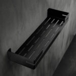 Czarna, metalowa półka łazienkowa VEGA z matowym wykończeniem, zamontowana na ścianie, z widocznymi kroplami wody. Na półce ustawione są eleganckie kosmetyki w ciemnych butelkach. Konstrukcja posiada perforowane dno dla odpływu wody oraz wysokie boki zabezpieczające przed zsuwaniem się przedmiotów.