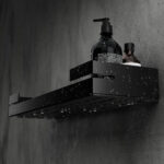 Czarna, metalowa półka łazienkowa VEGA z matowym wykończeniem, zamontowana na ścianie, z widocznymi kroplami wody. Na półce ustawione są eleganckie kosmetyki w ciemnych butelkach. Konstrukcja posiada perforowane dno dla odpływu wody oraz wysokie boki zabezpieczające przed zsuwaniem się przedmiotów.