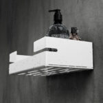 Biała, metalowa półka łazienkowa VEGA z matowym wykończeniem, zamontowana na ścianie, z widocznymi kroplami wody. Na półce ustawione są eleganckie kosmetyki w ciemnych butelkach. Konstrukcja posiada perforowane dno dla odpływu wody oraz wysokie boki zabezpieczające przed zsuwaniem się przedmiotów.