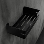 Czarna, metalowa półka łazienkowa VEGA z matowym wykończeniem, zamontowana na ścianie, z widocznymi kroplami wody. Na półce ustawione są eleganckie kosmetyki w ciemnych butelkach. Konstrukcja posiada perforowane dno dla odpływu wody oraz wysokie boki zabezpieczające przed zsuwaniem się przedmiotów.