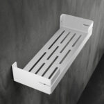 Biała, metalowa półka łazienkowa VEGA z matowym wykończeniem, zamontowana na ścianie, z widocznymi kroplami wody. Na półce ustawione są eleganckie kosmetyki w ciemnych butelkach. Konstrukcja posiada perforowane dno dla odpływu wody oraz wysokie boki zabezpieczające przed zsuwaniem się przedmiotów.