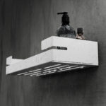 Biała, metalowa półka łazienkowa VEGA z matowym wykończeniem, zamontowana na ścianie, z widocznymi kroplami wody. Na półce ustawione są eleganckie kosmetyki w ciemnych butelkach. Konstrukcja posiada perforowane dno dla odpływu wody oraz wysokie boki zabezpieczające przed zsuwaniem się przedmiotów.