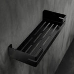 Czarna, metalowa półka łazienkowa VEGA z matowym wykończeniem, zamontowana na ścianie, z widocznymi kroplami wody. Na półce ustawione są eleganckie kosmetyki w ciemnych butelkach. Konstrukcja posiada perforowane dno dla odpływu wody oraz wysokie boki zabezpieczające przed zsuwaniem się przedmiotów.