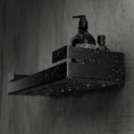 Czarna, metalowa półka łazienkowa VEGA z matowym wykończeniem, zamontowana na ścianie, z widocznymi kroplami wody. Na półce ustawione są eleganckie kosmetyki w ciemnych butelkach. Konstrukcja posiada perforowane dno dla odpływu wody oraz wysokie boki zabezpieczające przed zsuwaniem się przedmiotów.